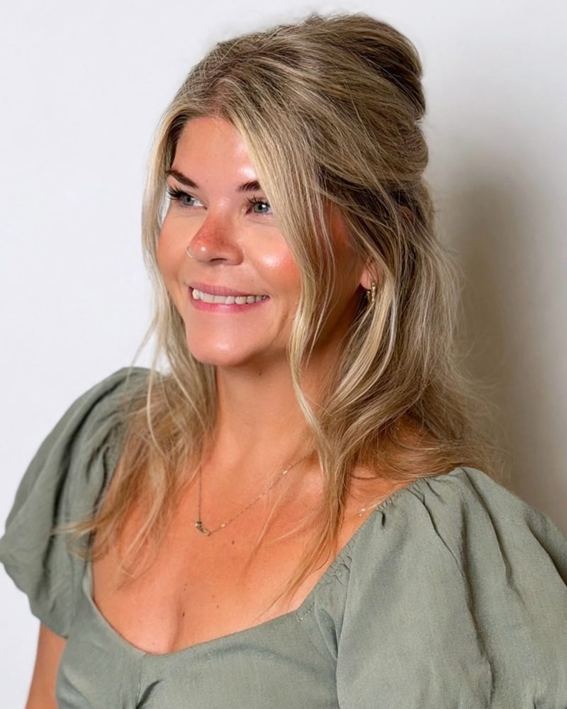 Melinda, owner of Colour Play Salon, smiling in a sage green puff-sleeve dress with her blonde hair in a half-up style
